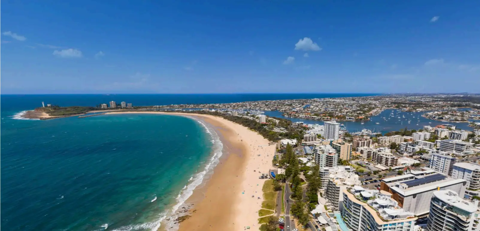 Sunshine-coast-image | Compass Transport Aerial view of a curved sandy beach along a turquoise bay, with a dense city of high-rise buildings along the shore and a marina on the right.